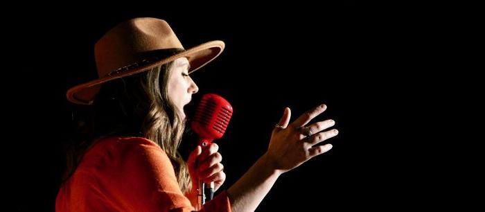 A person wearing a tan hat and an orange shirt holds a red vintage-style microphone and gestures with their hand while singing against a black background.